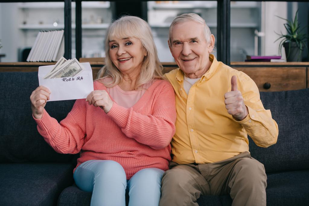 Seniorenpaar zeigt Daumen hoch, während es einen Umschlag mit "roth ira" -Schriftzug und Dollarnoten in der Hand hält - Foto, Bild