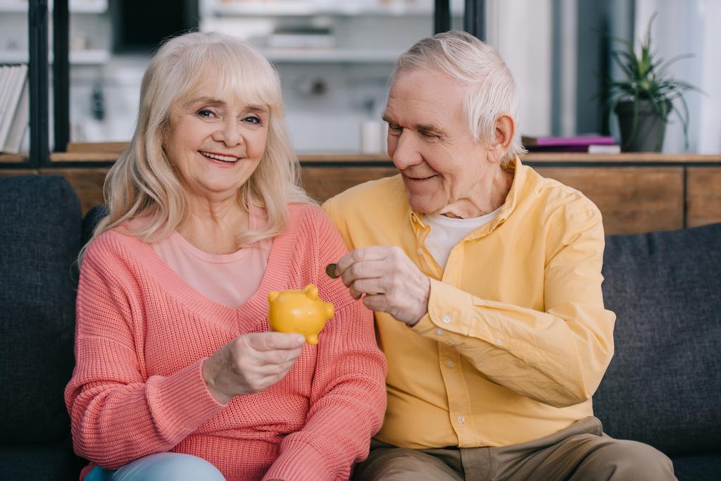 Glückliches Seniorenpaar steckt Münze in gelbes Sparschwein - Foto, Bild