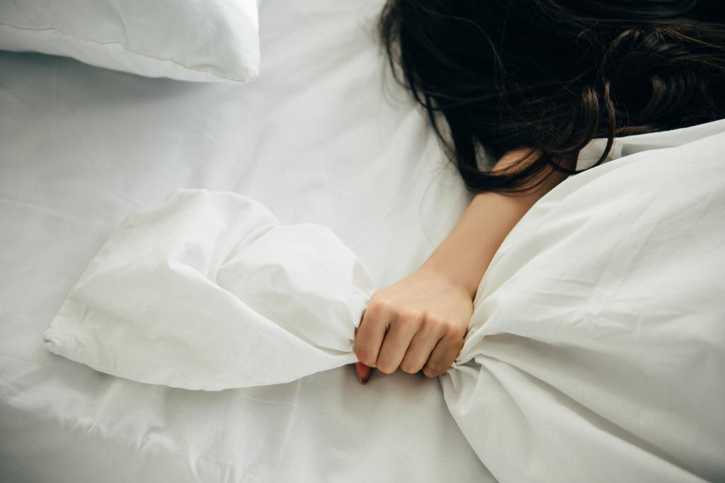 Ausgeschnittene Ansicht einer Frau, die weißes Bettzeug in der Hand hält, während sie auf dem Bett liegt - Foto, Bild