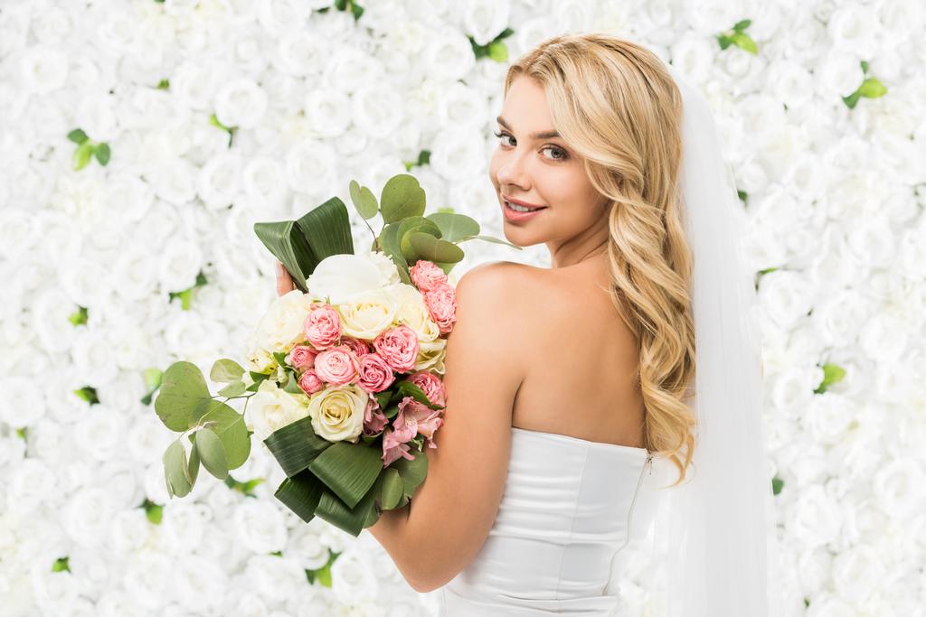 mooie jonge bruid houden bruiloft boeket en camera kijken op witte bloemen achtergrond - Foto, afbeelding