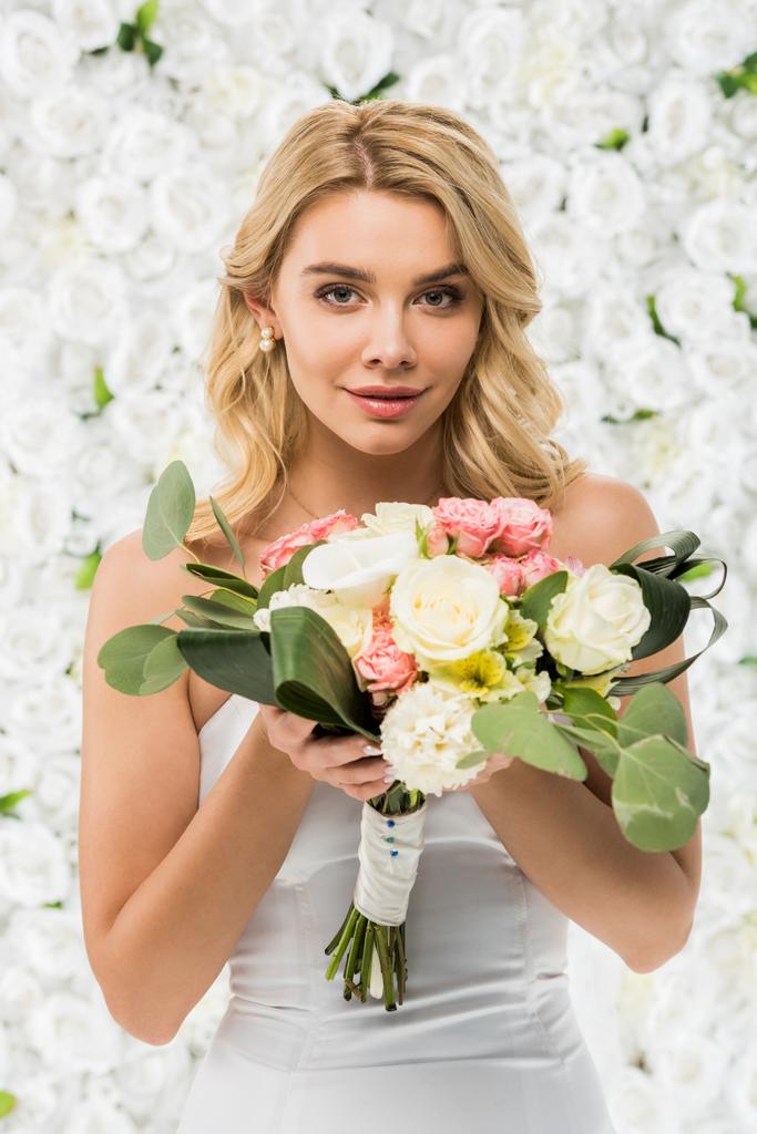 красивая молодая женщина держит свадебный букет и смотрит в камеру на белом цветочном фоне
 - Фото, изображение