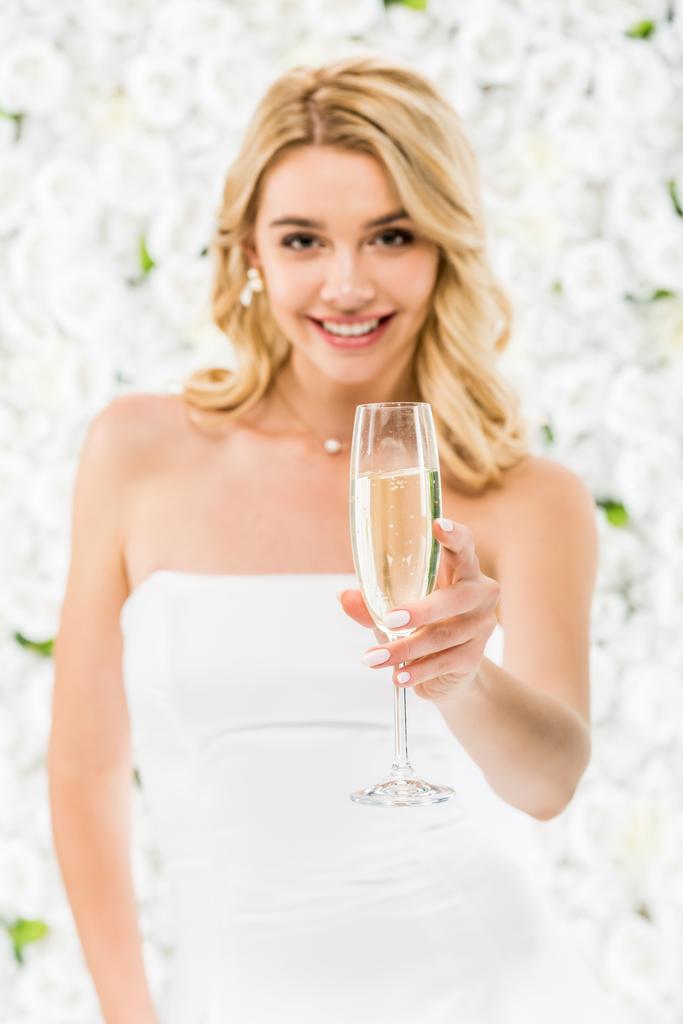 selective focus of smiling beautiful bride holding glass of champagne on white floral background - Photo, Image