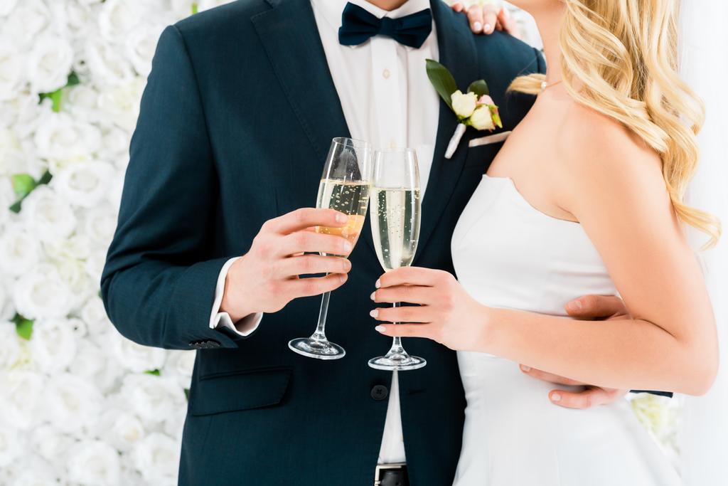 abgeschnittene Ansicht des Bräutigams und der Braut mit Champagnergläsern auf weißem Blumenhintergrund - Foto, Bild