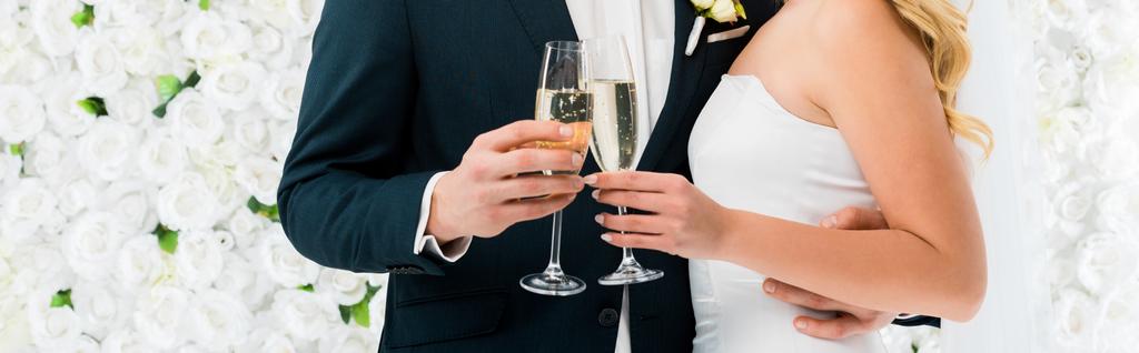 damat ve gelin bardak şampanya beyaz çiçek arka plan üzerinde tutarak panoramik çekim - Fotoğraf, Görsel
