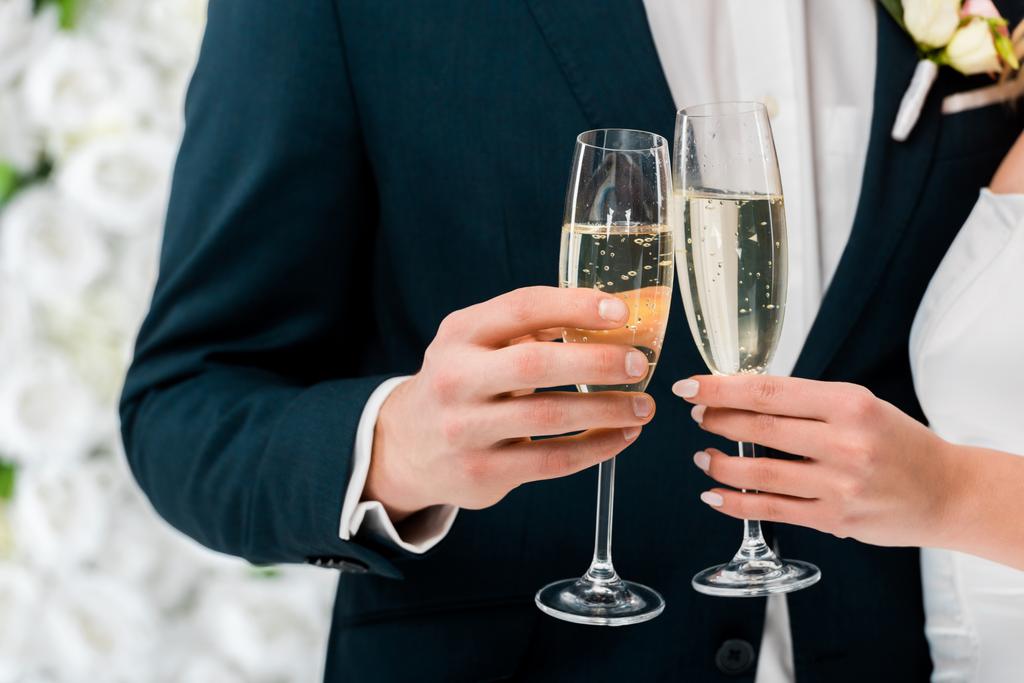 Vista parcial del novio y la novia con copas de champán sobre fondo floral blanco
 - Foto, Imagen