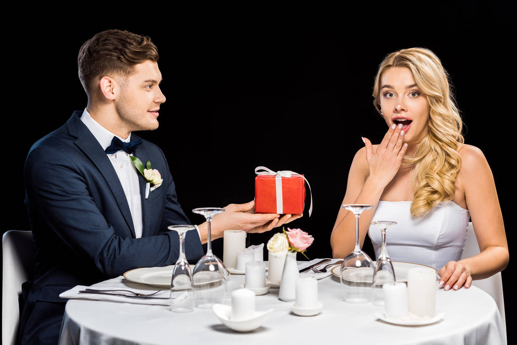 Lächelnder junger Bräutigam überreicht rote Geschenkschachtel an überraschte Braut isoliert auf Schwarz - Foto, Bild