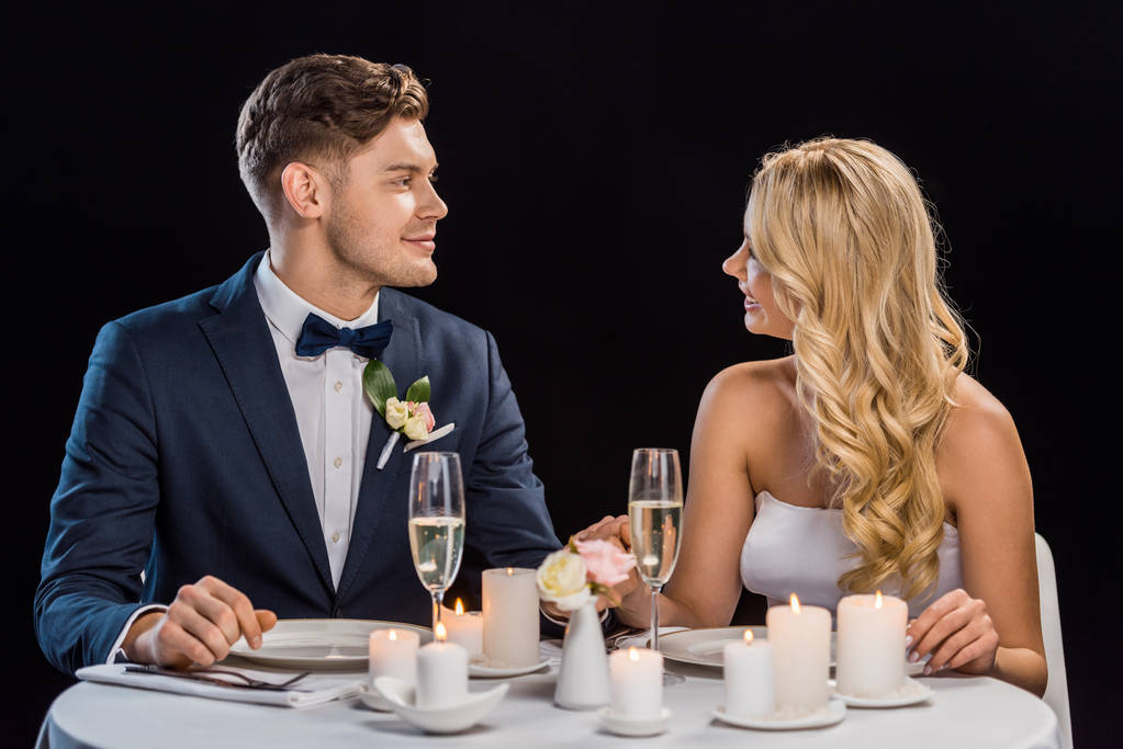 feliz joven pareja sentado a la mesa servida y mirándose el uno al otro aislado en negro
 - Foto, Imagen