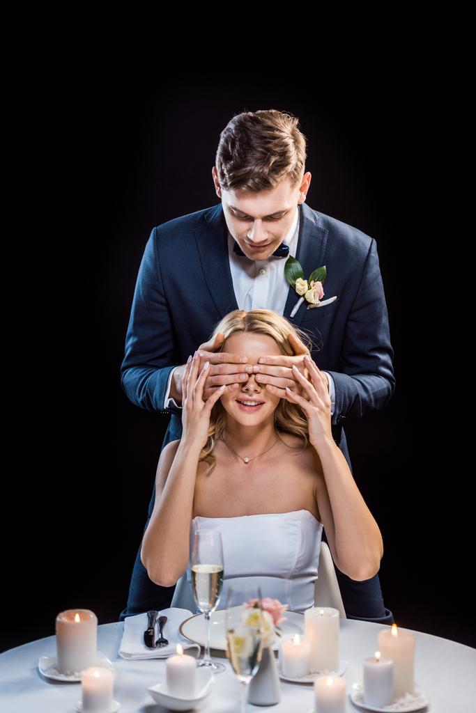 young groom standing behind sitting bride and closing her eyes with hands isolated on black - Photo, Image