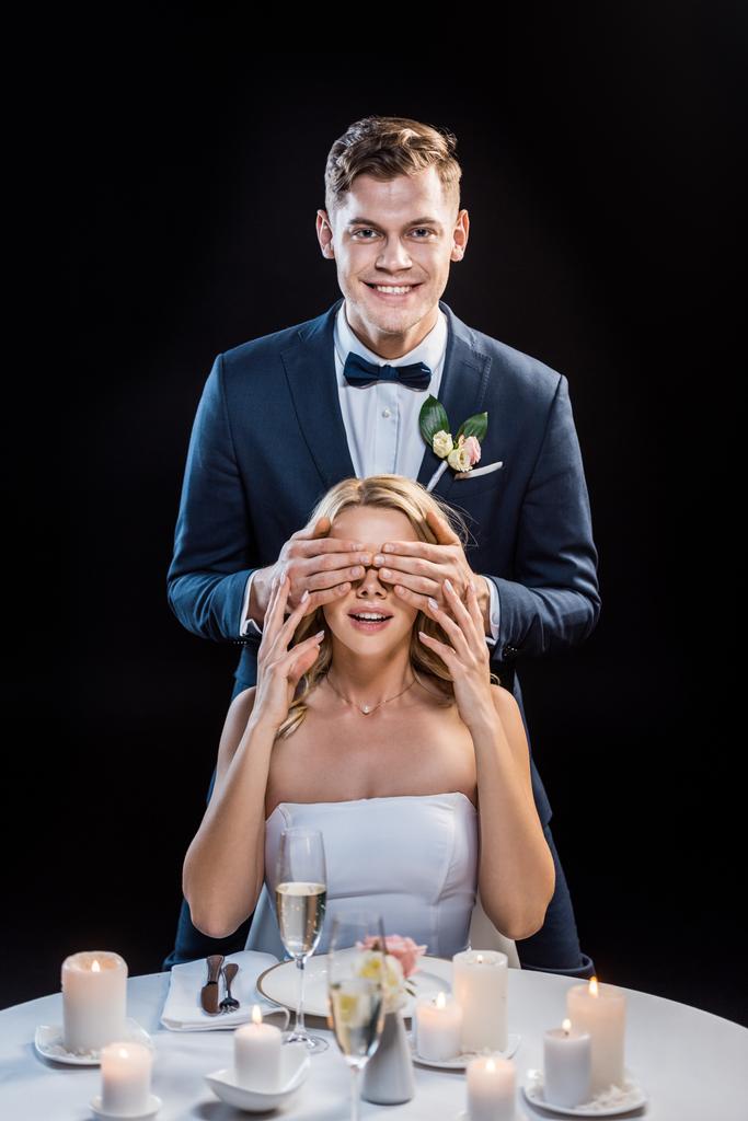 glücklicher Bräutigam, der hinter der lächelnden Braut steht und ihre Augen schließt, die Hände isoliert auf schwarz - Foto, Bild