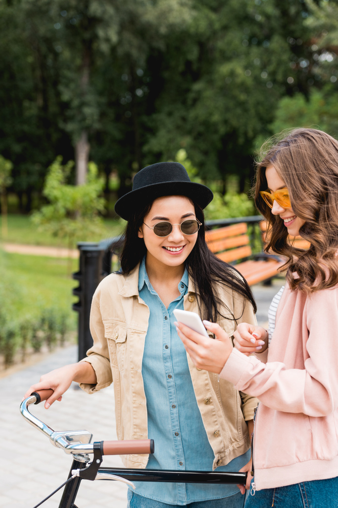 happy girls in sunglasses smiling while looking at smartphone and standing near bike in park - Photo, Image