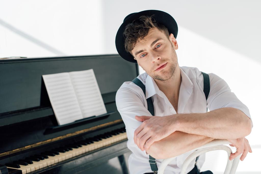 zasněný pianista v bílé košili a černém klobouku při pohledu na fotoaparát - Fotografie, Obrázek