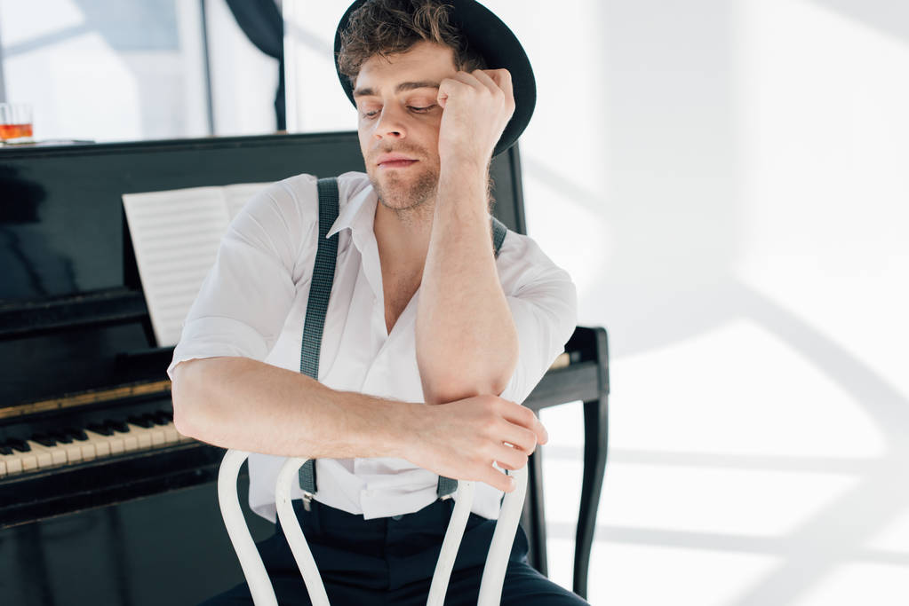 handsome dreamy pianist in white shirt and black hat sitting on white chair - Photo, Image