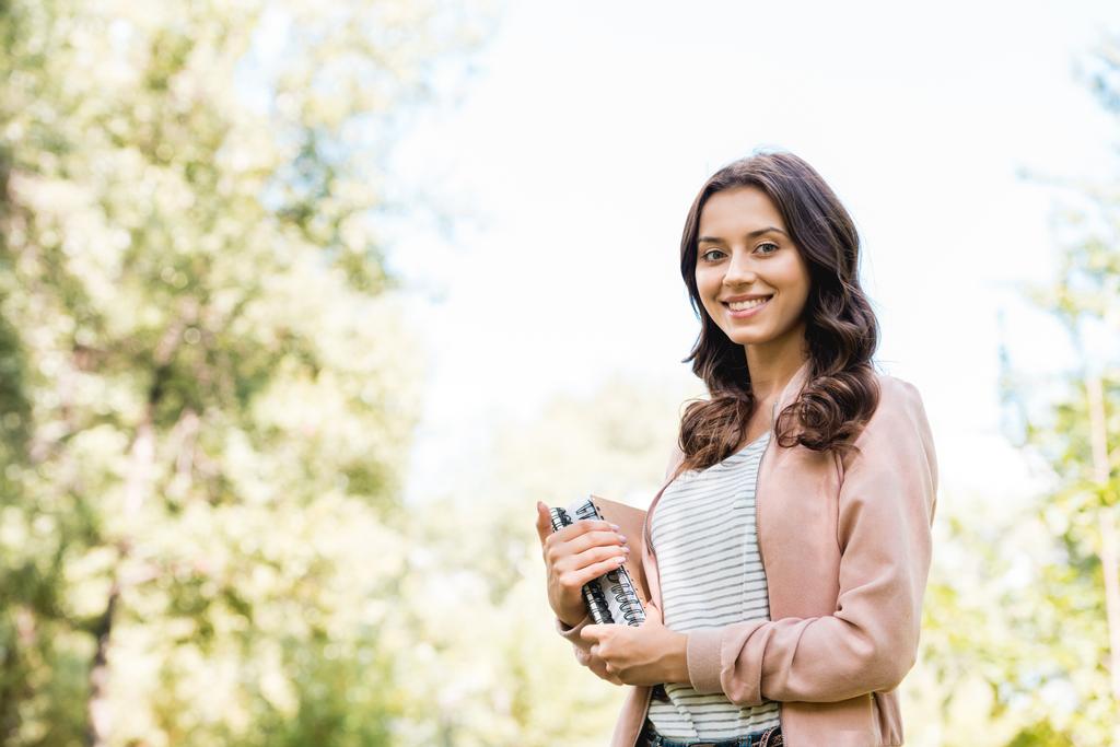 piękna młoda kobieta trzymania książek i uśmiechając się w parku - Zdjęcie, obraz