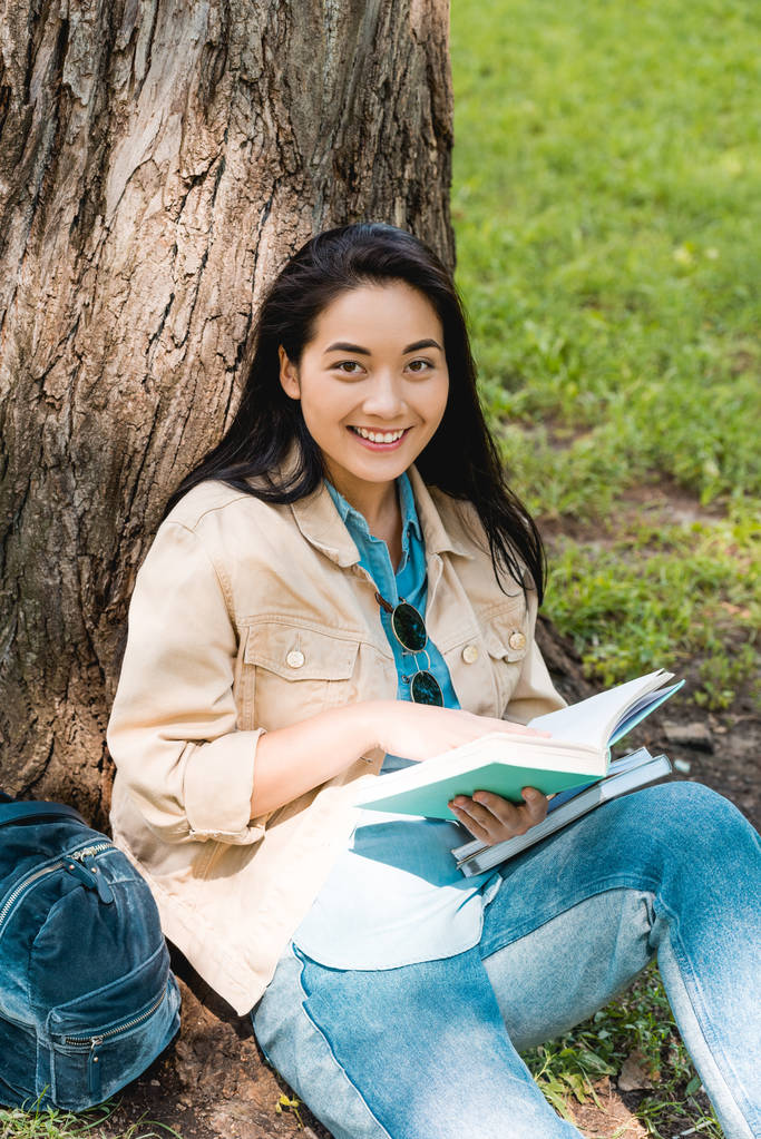 pretty student smiling while sitting in park with books - Photo, Image