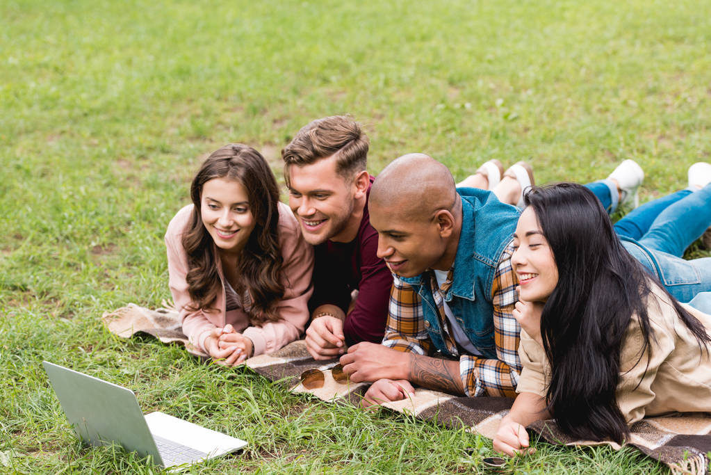 щасливі мультикультурні друзі лежать на ковдрі і дивляться на ноутбук у парку
 - Фото, зображення