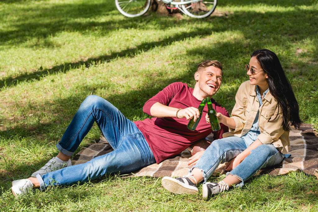 ευτυχισμένο ζευγάρι τσούγκριζαν μπουκάλια με μπύρα, ενώ κάθεται σε κουβέρτα στο πάρκο - Φωτογραφία, εικόνα