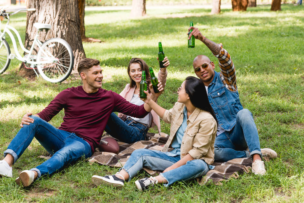 fröhliche multiethnische Gruppe von Freunden, die auf karierter Decke im Park sitzen und Flaschen klirren - Foto, Bild