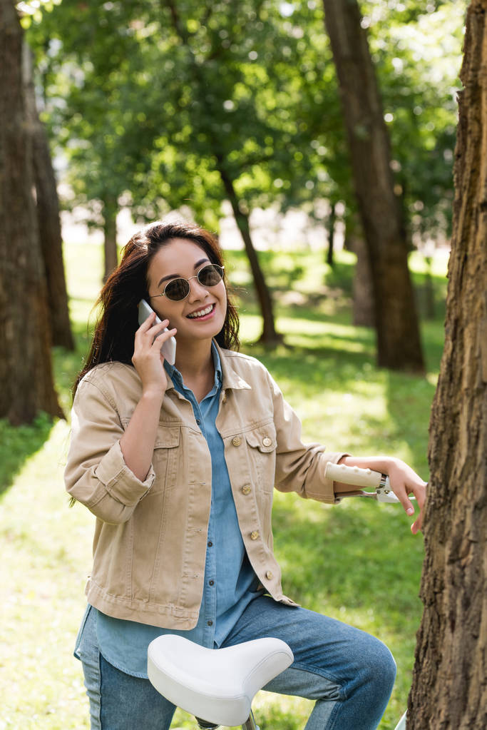 Gelukkig jonge vrouw in zonnebril praten over smartphone in de buurt van fiets in park - Foto, afbeelding