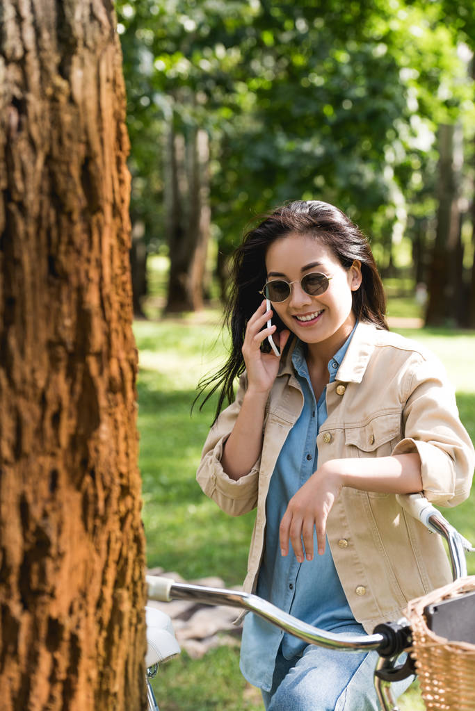 χαμογελώντας νεαρή γυναίκα σε γυαλιά ηλίου, μιλάμε για το smartphone κοντά ποδήλατο στο πάρκο - Φωτογραφία, εικόνα