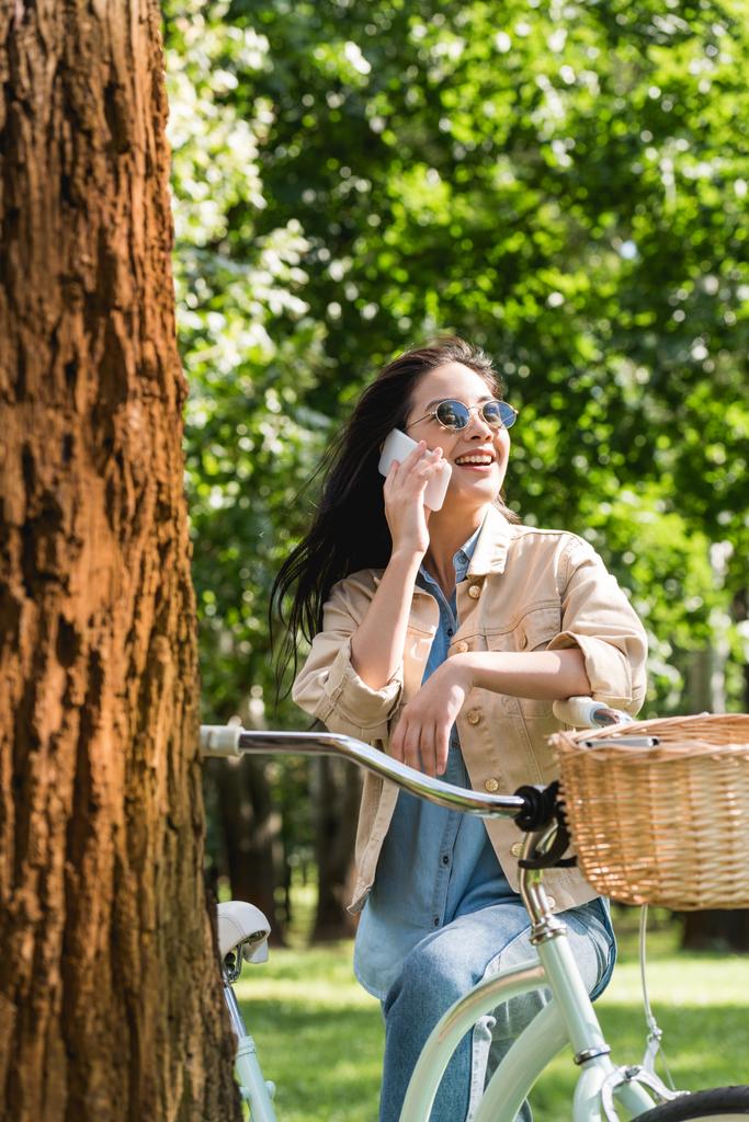 веселая девушка в солнечных очках разговаривает на смартфоне возле велосипеда в парке
 - Фото, изображение