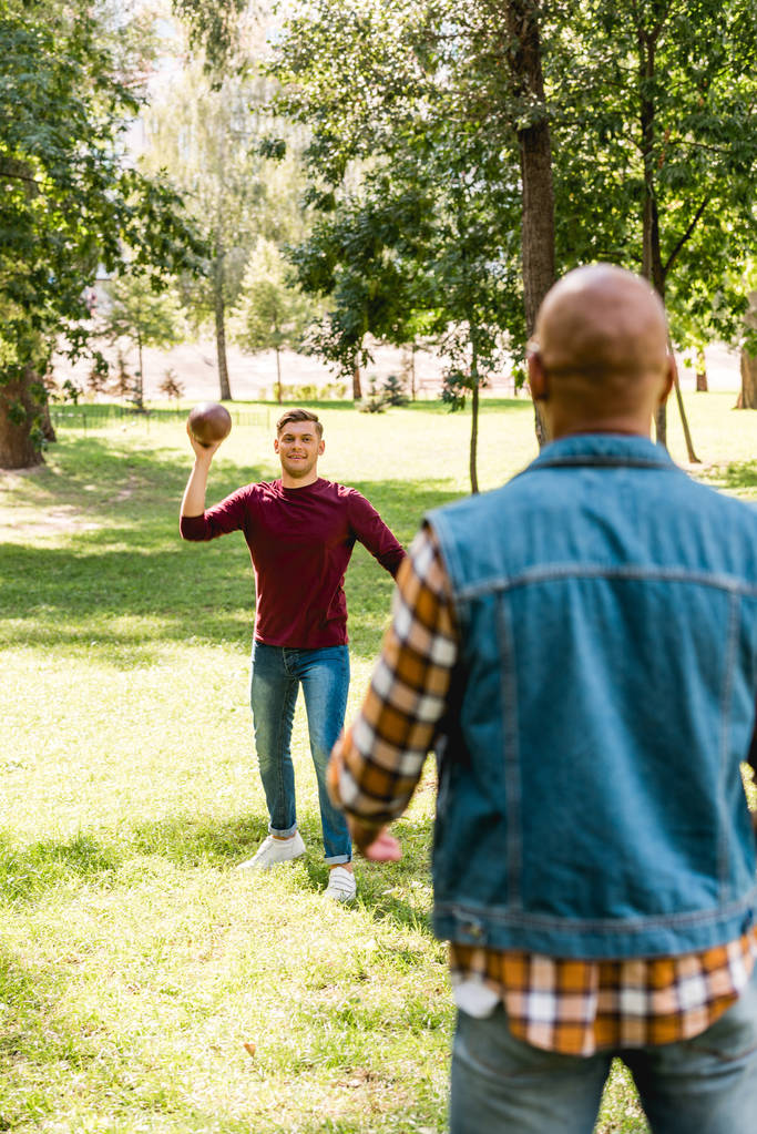 back view of african american man looking at happy friend throwing american football in park - Photo, Image