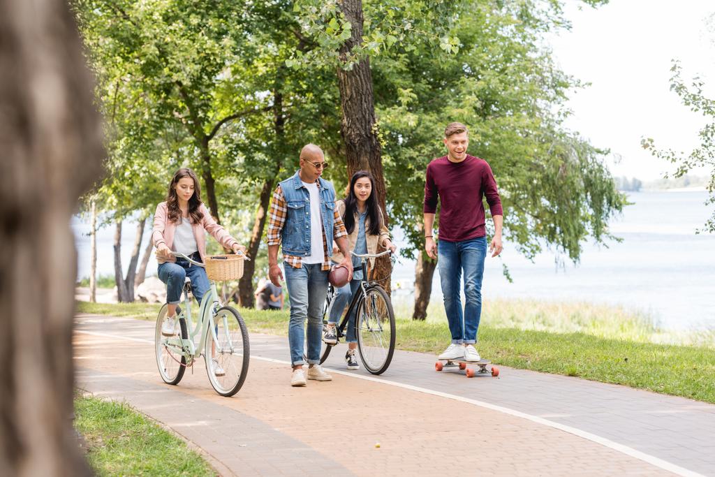 アフリカ系アメリカ人の女の子は、乗馬、自転車や公園内の陽気な友人ロングボーディング近くを歩いて  - 写真・画像
