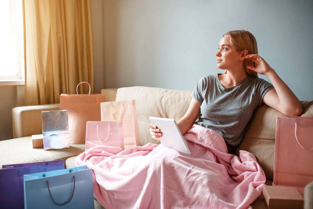 Compras online em casa. Jovem loira com tablet está pensando em mercadorias enquanto se senta com cobertor em um sofá
 - Foto, Imagem