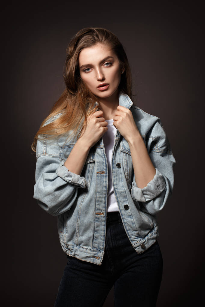 Gorgeous brunette girl with long flowing hair dressed in jeans jacket and jeans poses standing on the dark background in the studio - Photo, Image