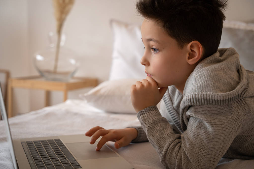 Um menino sentado em uma cama grande com um livro de laptop e olha para a câmera
. - Foto, Imagem
