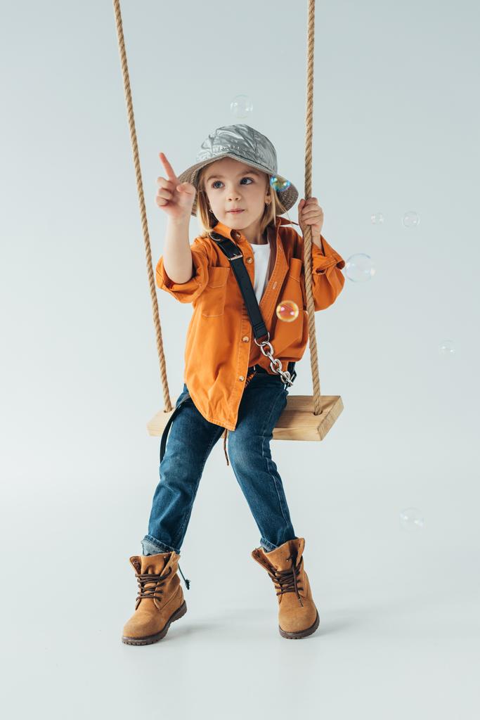 cute kid in jeans and orange shirt sitting on swing and ponting with finger at soap bubbles - Photo, Image