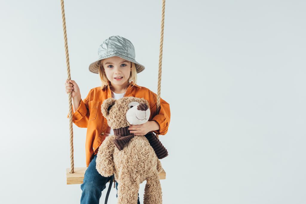 χαριτωμένο παιδί κάθεται στο swing και κρατώντας αρκουδάκι που απομονώνονται σε γκρι  - Φωτογραφία, εικόνα