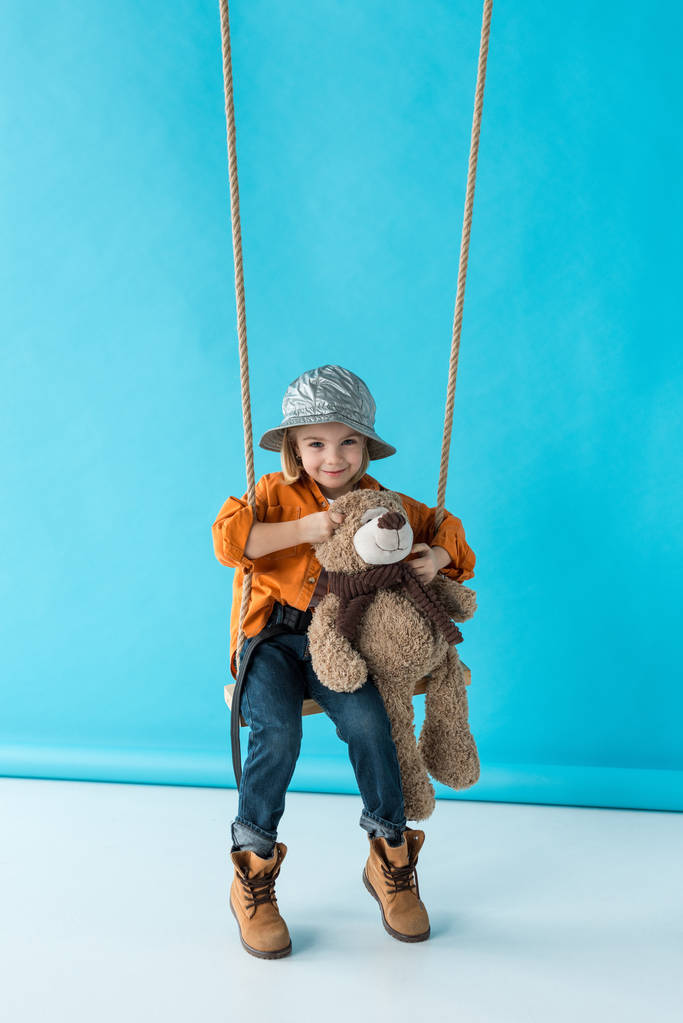 niedliches Kind sitzt auf Schaukel und hält Teddybär auf blauem Hintergrund  - Foto, Bild