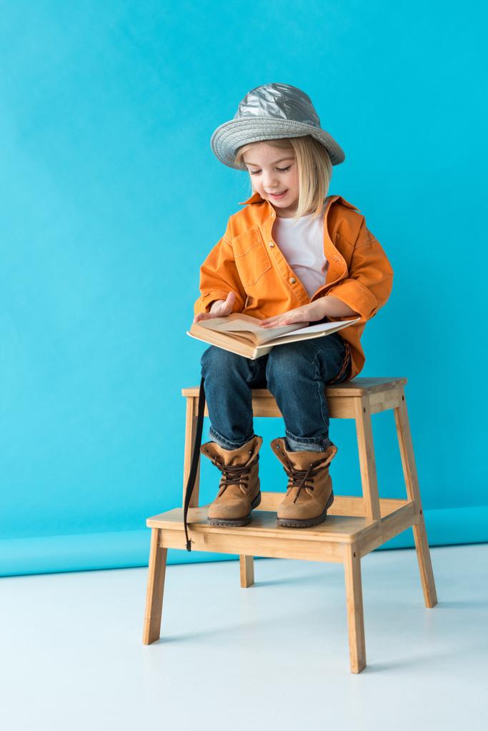 kid in jeans and orange shirt sitting on stairs and reading book on blue background  - Photo, Image