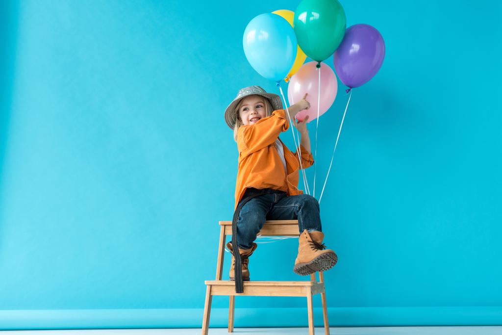 cute kid in jeans and orange shirt sitting on stairs and pointing with finger at balloons - Photo, Image