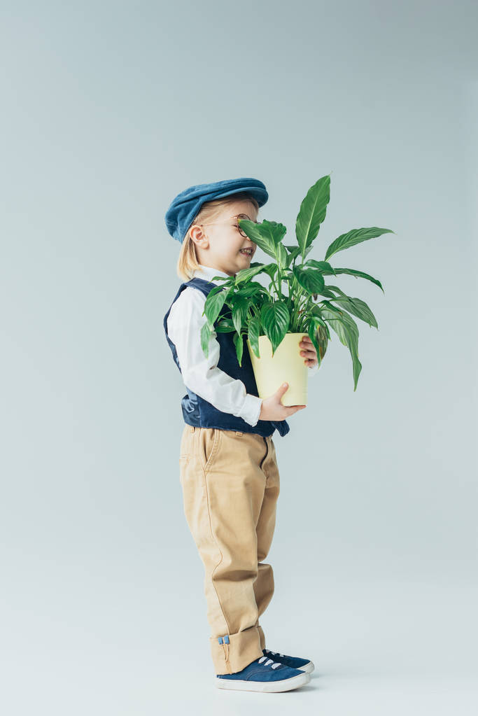 entzückendes Kind in Retro-Weste und Mütze mit Pflanze im Blumentopf auf grauem Hintergrund  - Foto, Bild
