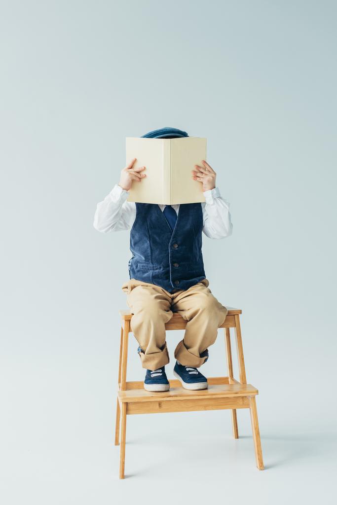 Kid zittend op houten trappen en lezen boek over grijze achtergrond  - Foto, afbeelding