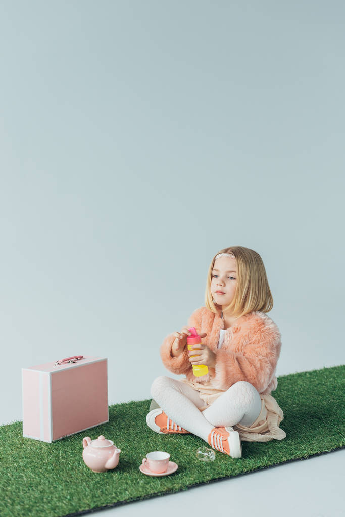 cute kid with crossed legs sitting on grass rug and holding bottle with soap bubbles isolated on grey - Photo, Image