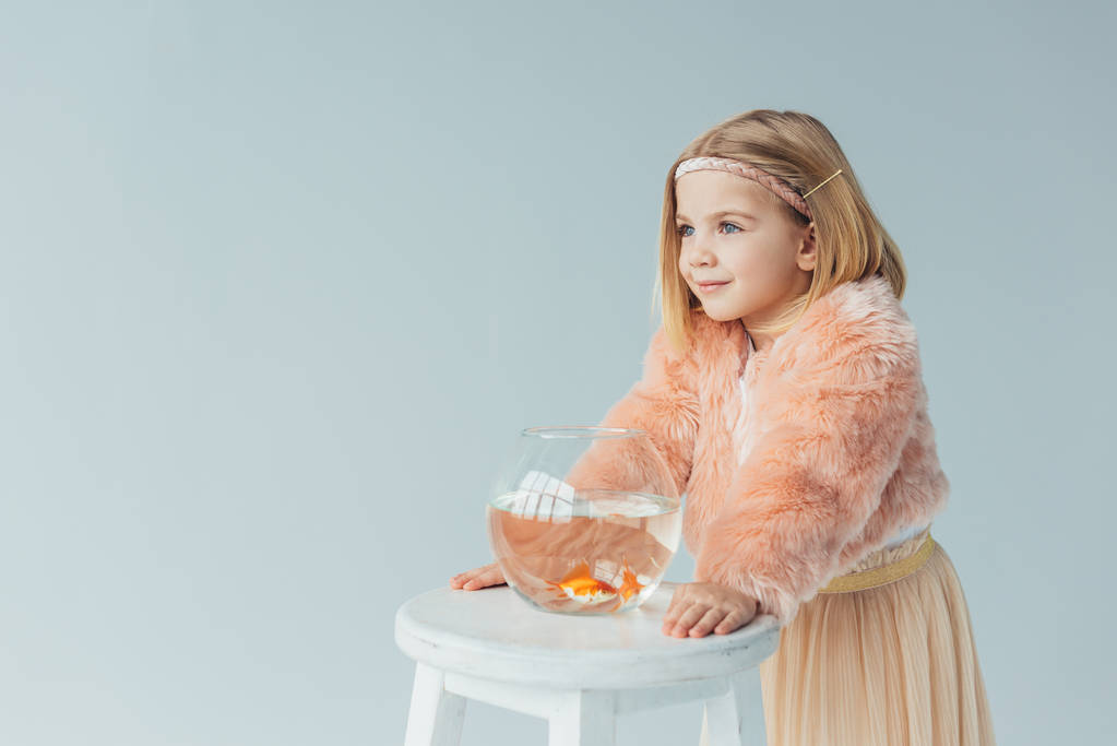 cute kid in faux fur coat and skirt with fishbowl looking away isolated on grey - Photo, Image