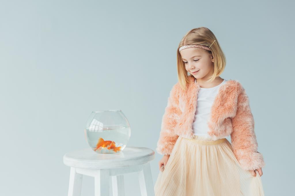 чарівна дитина в штучному шубці і спідниці, дивлячись на акваріум на табуреті ізольовані на сірому
 - Фото, зображення