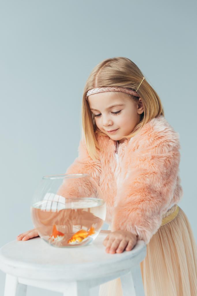adorable kid in faux fur coat and skirt looking at fishbowl on stool isolated on grey - Photo, Image
