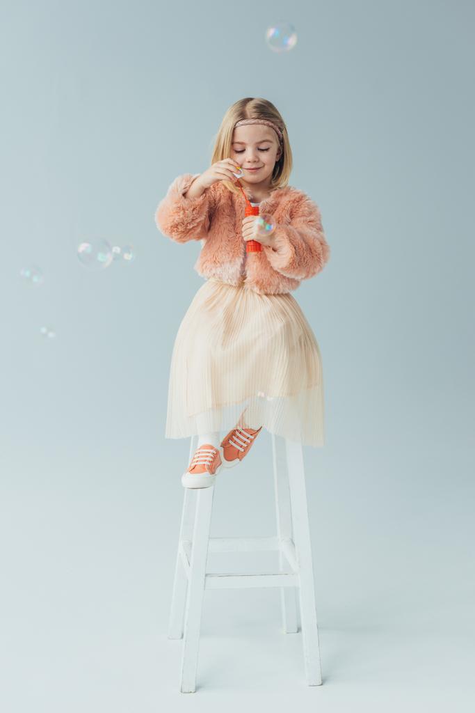 adorable kid in faux fur coat and skirt sitting on highchair and blowing soap bubbles - Photo, Image