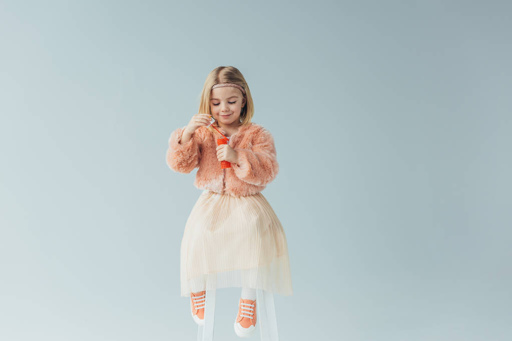 adorable kid in faux fur coat and skirt sitting on highchair and holding bottle with soap bubbles isolated on grey - Photo, Image