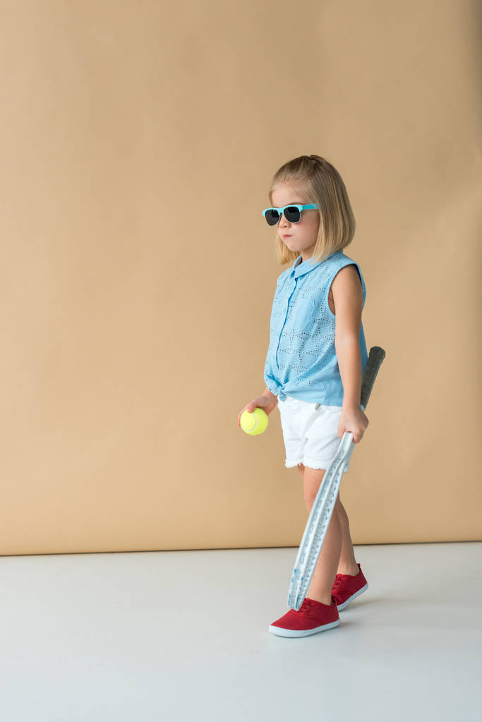 cute kid in sunglasses, shirt and shorts holding racket and ball on beige background  - Photo, Image