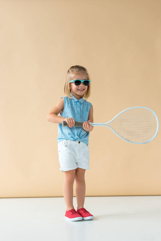усміхнена і мила дитина в сонцезахисних окулярах, сорочках і шортах тримає ракетку на бежевому фоні
  - Фото, зображення