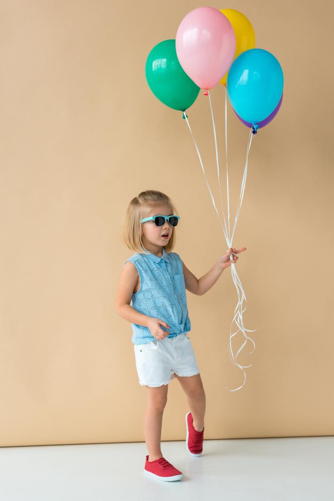 lindo niño en gafas de sol, camisa y pantalones cortos sosteniendo globos sobre fondo beige
  - Foto, imagen