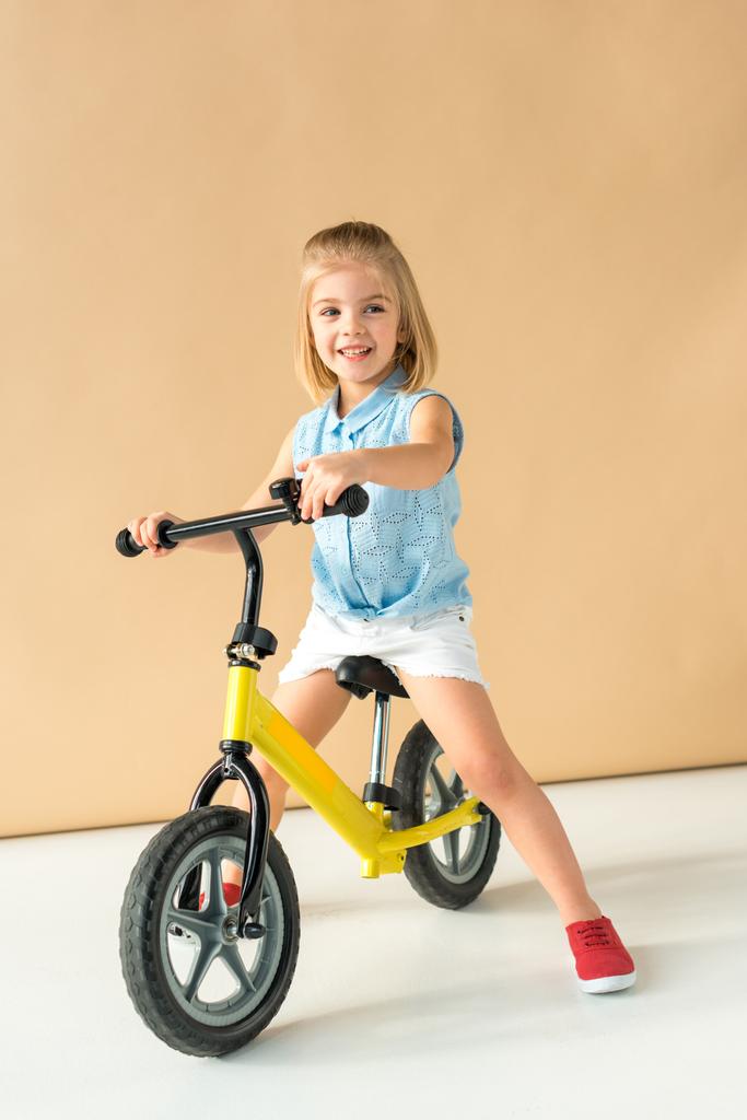 smiling kid in shirt and shorts riding bicycle on beige background  - Photo, Image