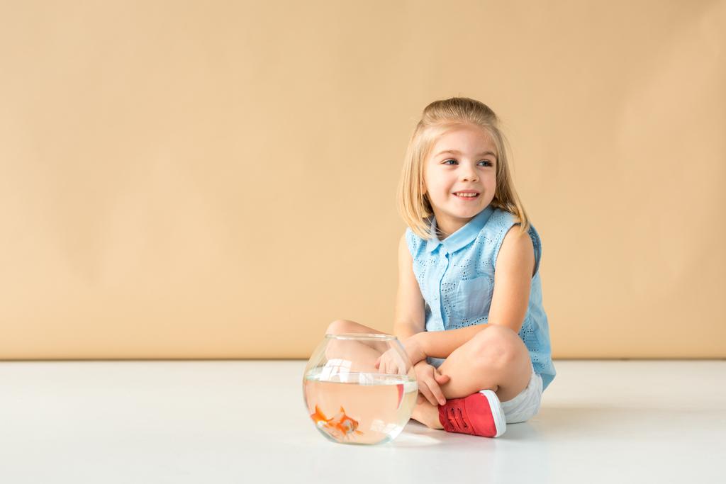 carino bambino seduto sul pavimento con pesce ciotola e guardando lontano su sfondo beige
  - Foto, immagini