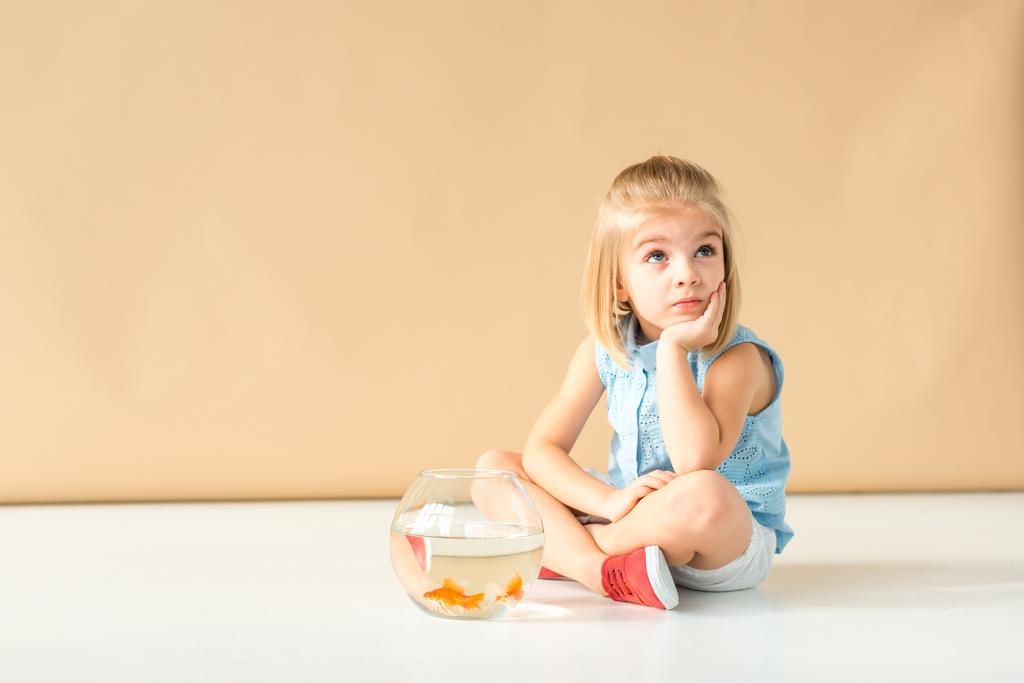 verträumtes Kind sitzt mit Fischschale auf dem Boden und schaut auf beigem Hintergrund weg  - Foto, Bild