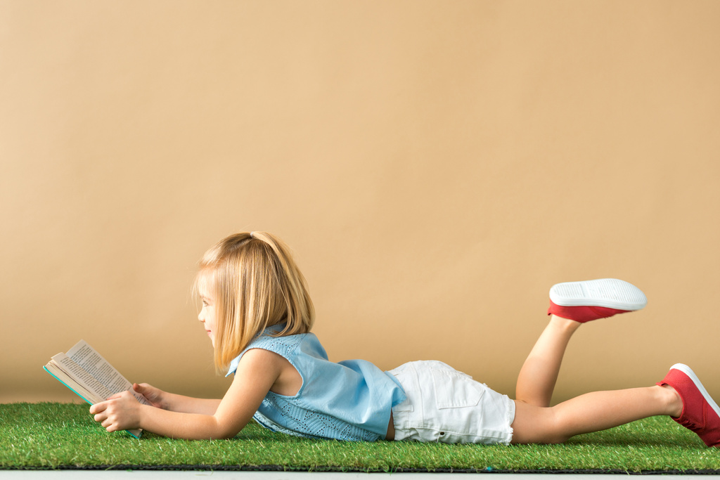 a gyerek feküdt szőnyeg fű és bézs háttérrel könyvével oldalnézete  - Fotó, kép