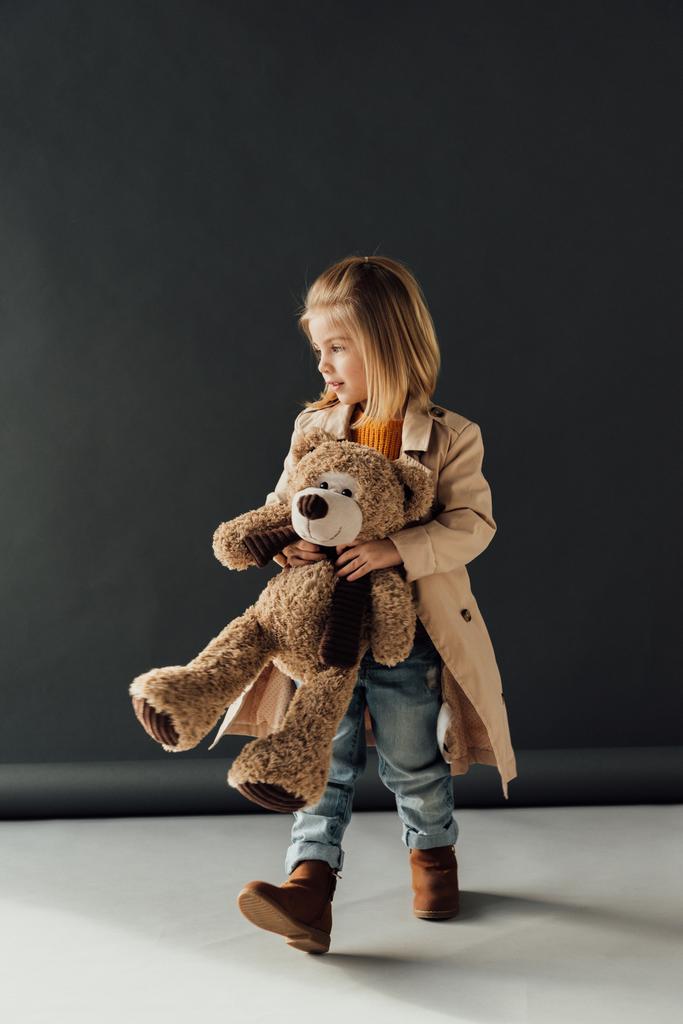 schattig kind in trenchcoat en jeans holding teddybeer op zwarte achtergrond - Foto, afbeelding